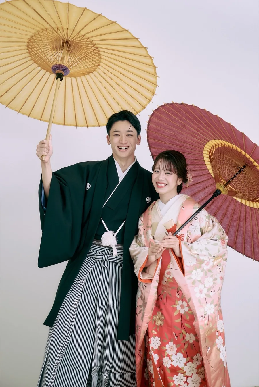 色打掛の花嫁と紋付袴の新郎が、和傘を手に笑顔で並ぶ婚礼写真